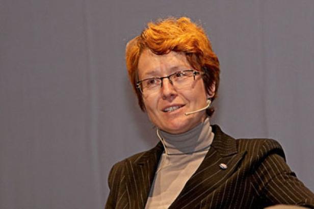 Eine Frau mit roten Haaren, Brille und Mikrofon spricht vor einem grauen Hintergrund.