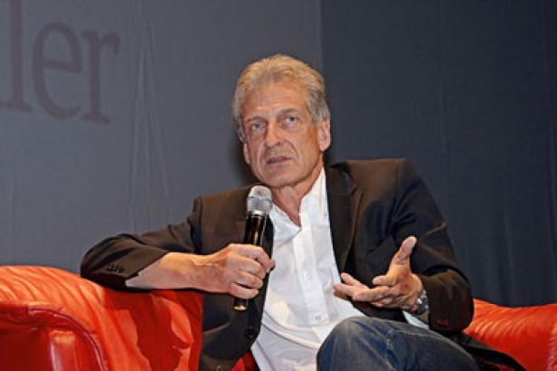 Ein Mann mit Mikrofon sitzt auf einem roten Sofa und gestikuliert.