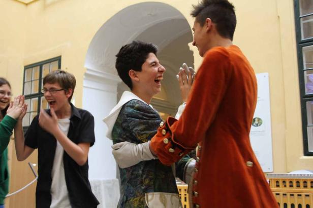 Zwei Jugendliche in historischen Kostümen lachen sich an, während andere applaudieren.