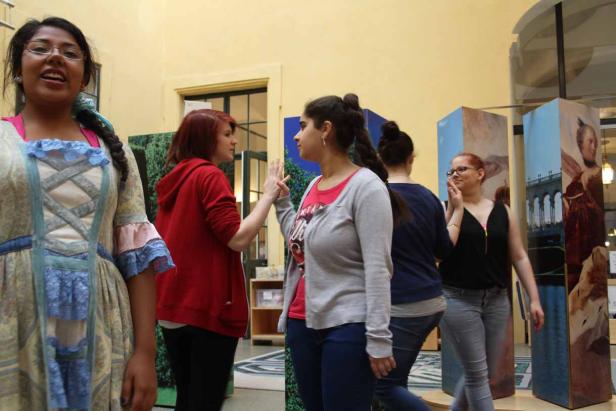 Mehrere junge Frauen stehen in einem Raum, einige in Kostümen, und interagieren miteinander.