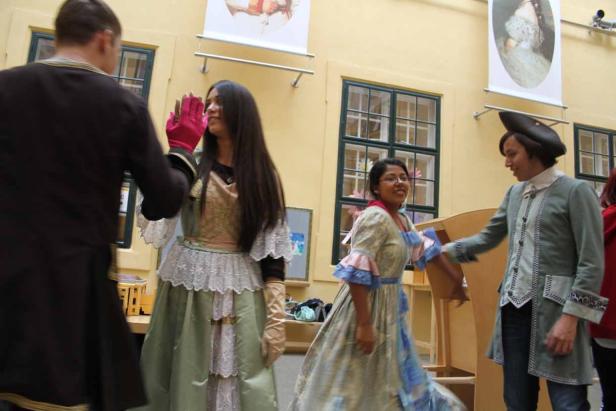 Personen in historischen Kostümen interagieren in einem Innenraum.