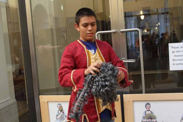 Ein junger Mann im historischen Kostüm hält eine graue Perücke in den Händen.
