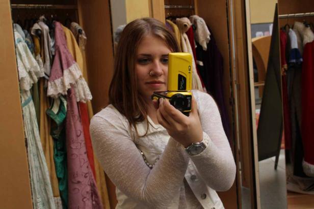 Eine junge Frau macht ein Selfie mit einer gelben Kamera vor einem Kleiderschrank voller Kostüme.