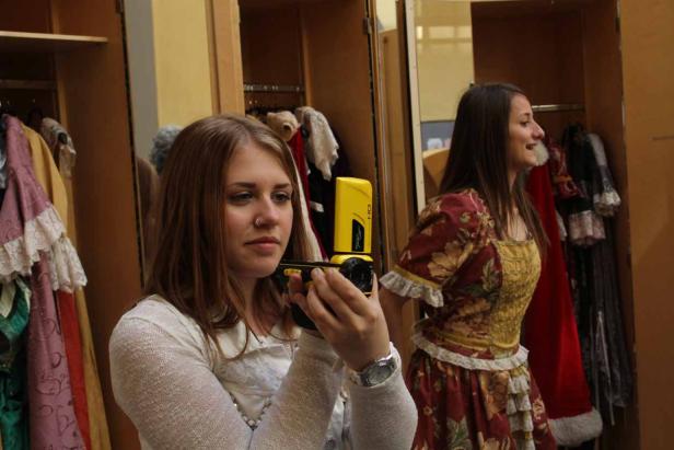 Eine junge Frau filmt mit einer gelben Kamera vor einem Kleiderschrank voller Kostüme.