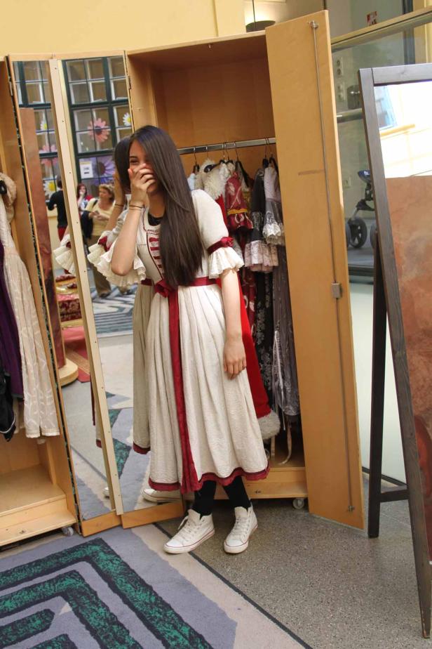 Eine junge Frau probiert ein historisch anmutendes Kleid an und lacht verlegen.