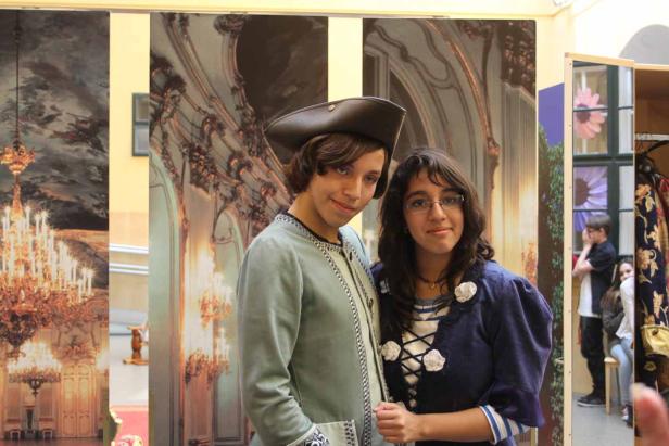 Ein junger Mann in historischer Kleidung und eine junge Frau posieren vor einer gemalten Palastkulisse.