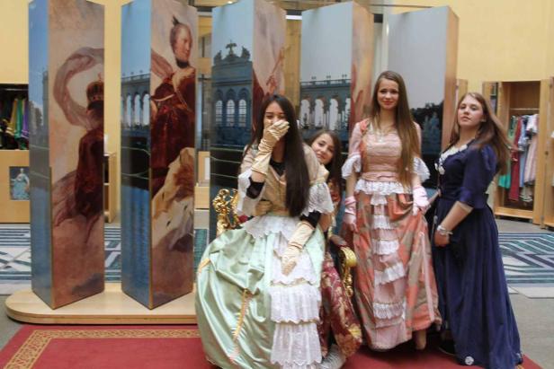 Vier junge Frauen posieren in historischen Kostümen vor bemalten Kulissen.