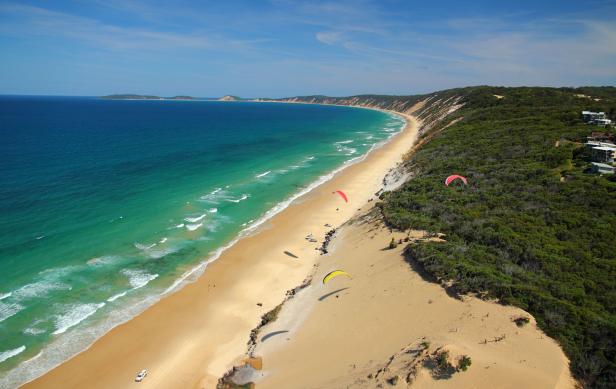 Ein Panoramablick auf einen langen Sandstrand mit türkisfarbenem Wasser und mehreren Gleitschirmfliegern.