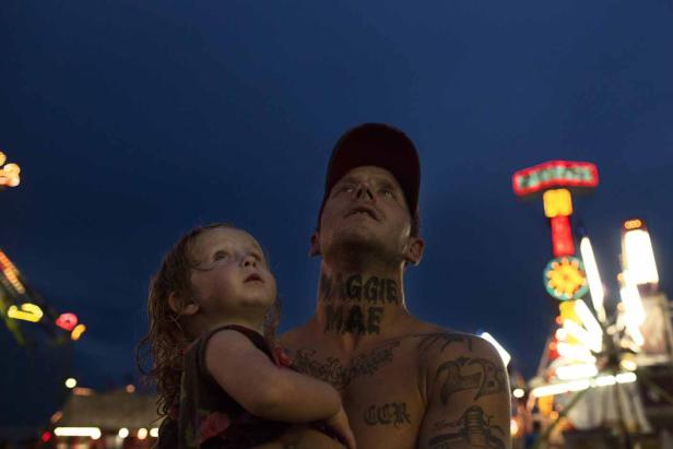 Ein Vater hält seine kleine Tochter auf einem Jahrmarkt im Arm, beide schauen nach oben.