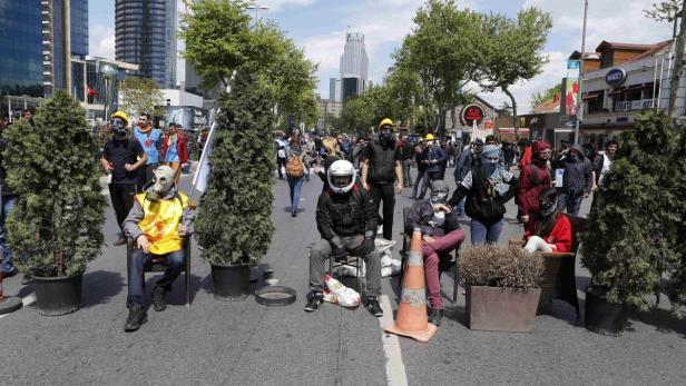 Eine Gruppe von Demonstranten sitzt mit Gasmasken und Helmen auf Stühlen auf einer belebten Straße.