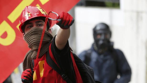 Ein Demonstrant mit rotem Helm und Schleuder im Vordergrund, eine weitere Person mit Gasmaske im Hintergrund.
