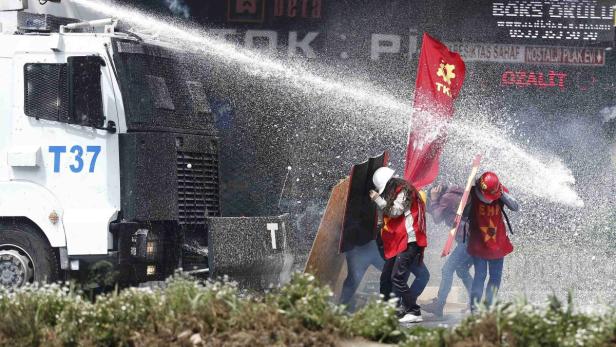 Ein Wasserwerfer sprüht auf Demonstranten mit roten Fahnen und Schutzschilden.