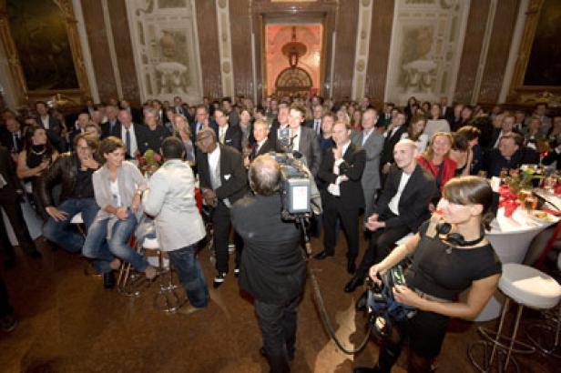 Eine Menschenmenge in einem eleganten Saal wird von einem Kamerateam gefilmt.