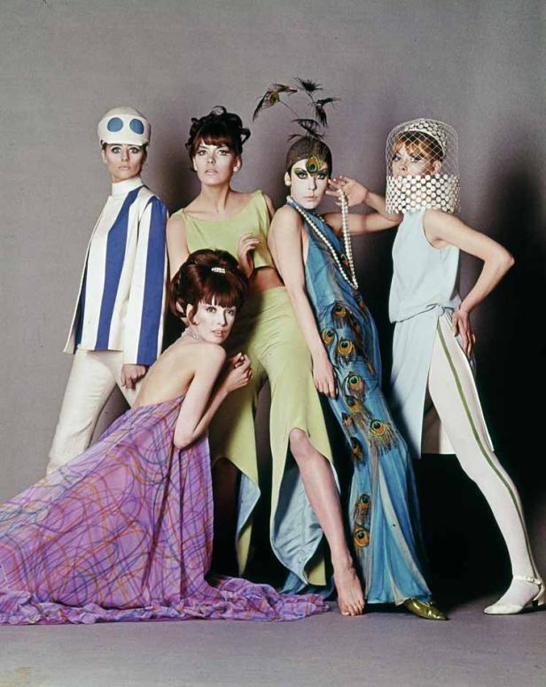 Fünf Models präsentieren farbenfrohe Mode und extravagante Kopfbedeckungen vor einem grauen Hintergrund.