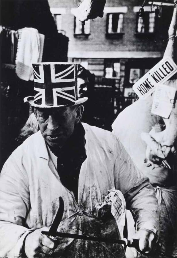 Ein Mann mit Union-Jack-Zylinder hantiert mit Werkzeugen vor einem Schild mit der Aufschrift „Home Killed“.