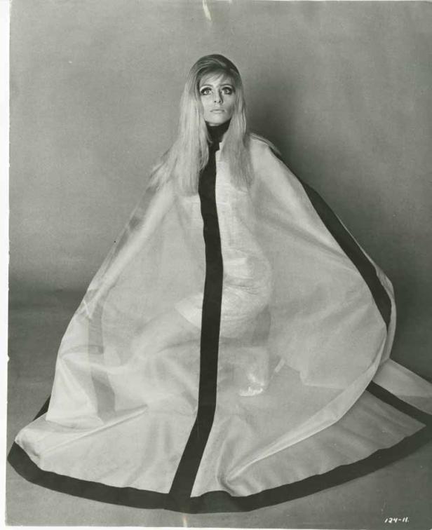 Eine Frau mit langen blonden Haaren trägt einen weißen Umhang mit schwarzem Besatz.