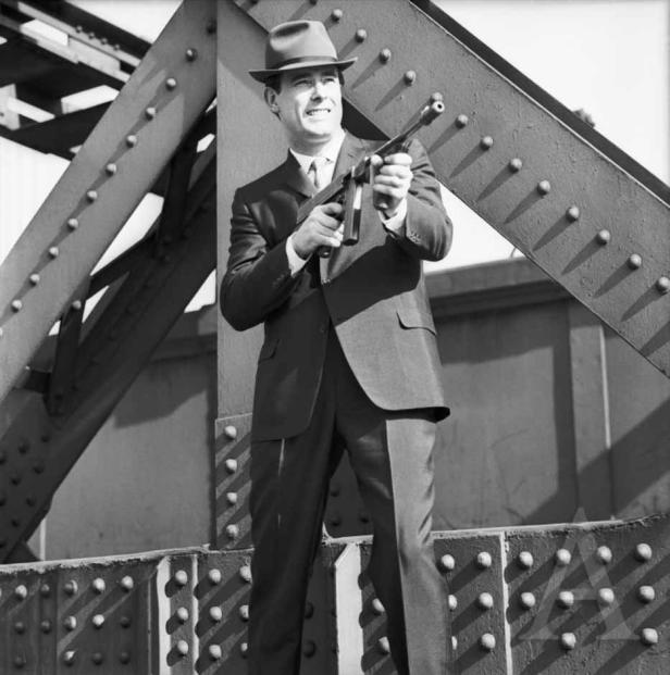 Ein Mann im Anzug und Hut steht auf einer Brücke und hält eine Maschinenpistole.