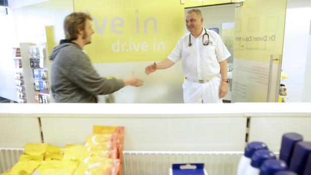 Ein Arzt im weißen Kittel begrüßt einen Patienten in einer Praxis mit der Aufschrift „drive in“.