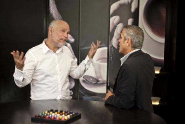 Nespresso/Episode 5/Making of/Mailand/George Clooney & John Malkovich (c: nespresso) Zwei Männer unterhalten sich an einem Tisch mit bunten Kugeln vor einer Kaffeetasse im Hintergrund.