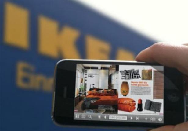 Eine Person fotografiert mit einem Smartphone eine IKEA-Katalogseite vor einem IKEA-Schild.