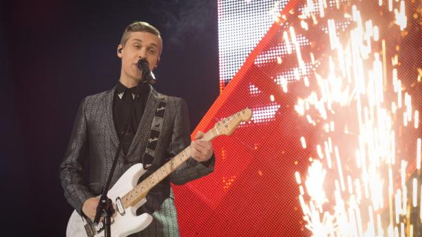 Ein Mann mit Gitarre singt vor einem roten Bildschirm mit Feuerwerk.