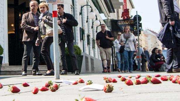 Auf einem Bürgersteig liegen Erdbeeren und Papier, während Passanten vorbeigehen.