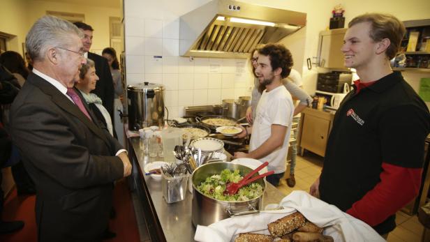 Ein Mann im Anzug besucht eine Küche, in der gekocht und Salat zubereitet wird.