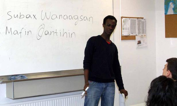 Ein junger Mann steht vor einer Tafel mit den Namen „Subax Wanaagsan“ und „Mafin Qantihin“.