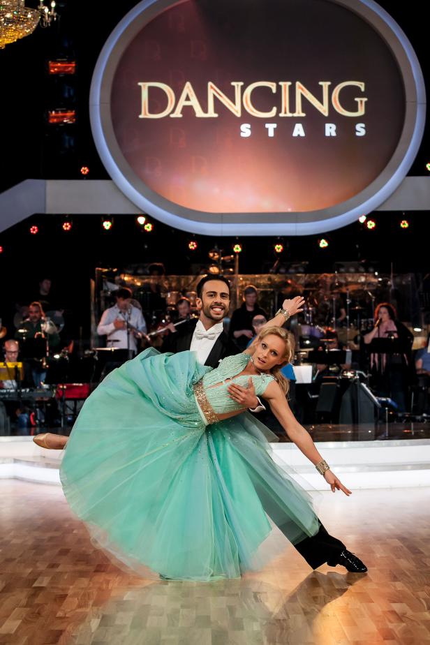 Ein Tanzpaar tanzt vor dem „Dancing Stars“-Logo mit einem Orchester im Hintergrund.