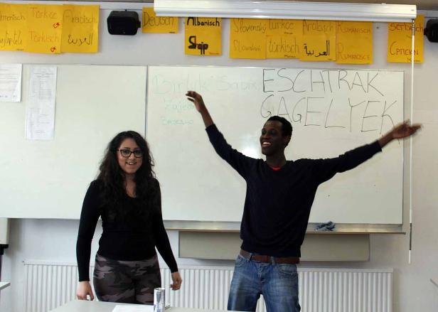 Eine Frau und ein Mann stehen vor einer Tafel mit Wörtern in verschiedenen Sprachen.