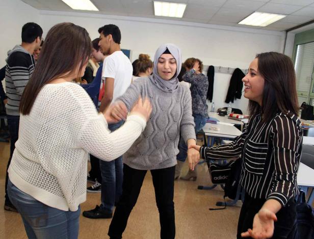 In einem Klassenzimmer schütteln sich zwei junge Frauen die Hände, während andere Schüler im Hintergrund stehen.