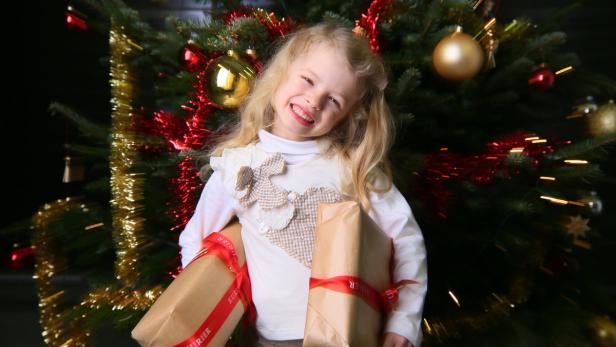 Ein lächelndes Mädchen hält Weihnachtsgeschenke vor einem geschmückten Weihnachtsbaum.