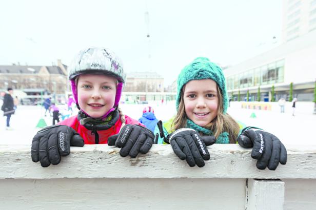 Zwei Mädchen schauen über eine hölzerne Bande auf einer Eisbahn.