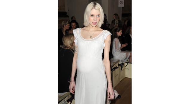 Eine schwangere Frau mit blonden Haaren und einem weißen Kleid sitzt bei einer Modenschau.
