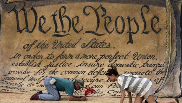 Zwei Personen knien vor einer Wand mit dem Text „We the People“ der US-Verfassung.