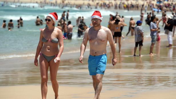 Ein Paar mit Weihnachtsmützen geht an einem sonnigen Strand entlang.