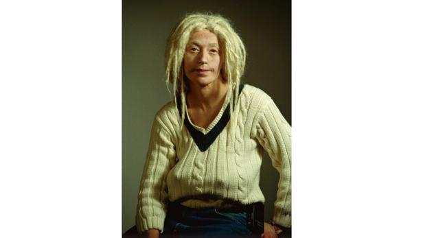 Eine Person mit blonden Dreads und einem cremefarbenen V-Ausschnitt-Pullover sitzt da.