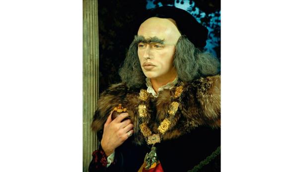 Ein Mann in historischer Kleidung mit Pelzkragen und auffälligen Augenbrauen hält eine Frucht.