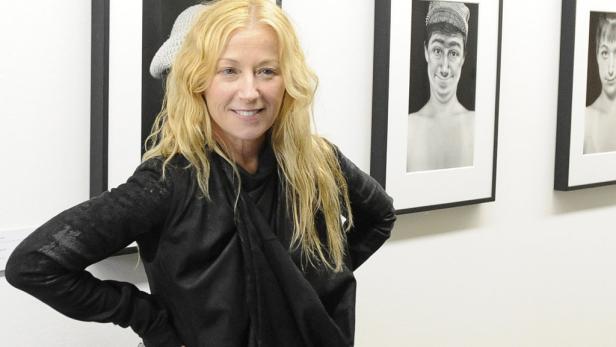 Eine Frau mit blonden Haaren steht vor einer Wand mit gerahmten Porträtzeichnungen.
