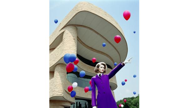 Eine Frau im lila Kostüm steht vor einem Gebäude, umgeben von roten und blauen Ballons.