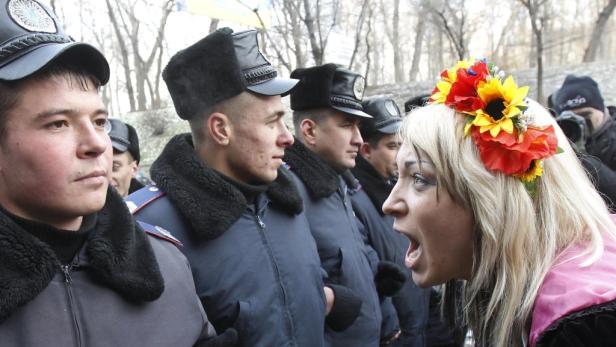 Eine Frau mit Blumenkranz schreit eine Reihe von uniformierten Männern an.