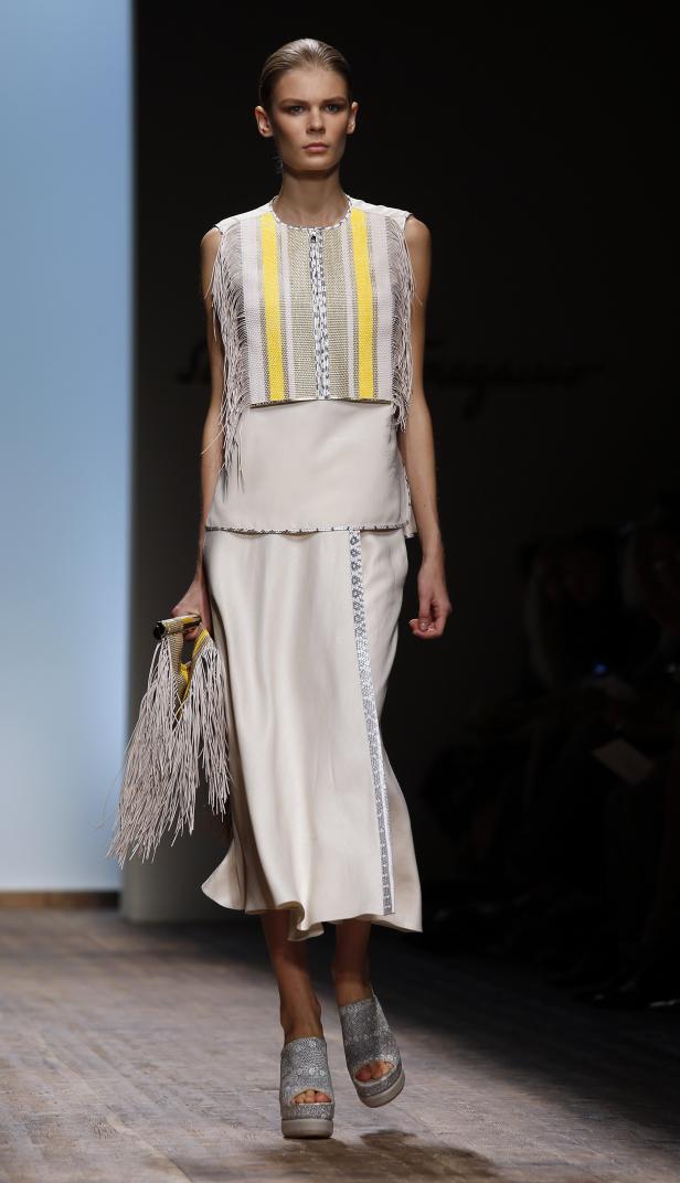 Ein Model präsentiert ein ärmelloses, helles Kleid mit Fransen und passender Handtasche auf dem Laufsteg.