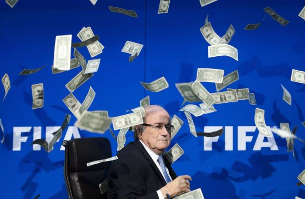 Sepp Blatter sitzt vor einem FIFA-Logo, während Geldscheine auf ihn herabregnen.