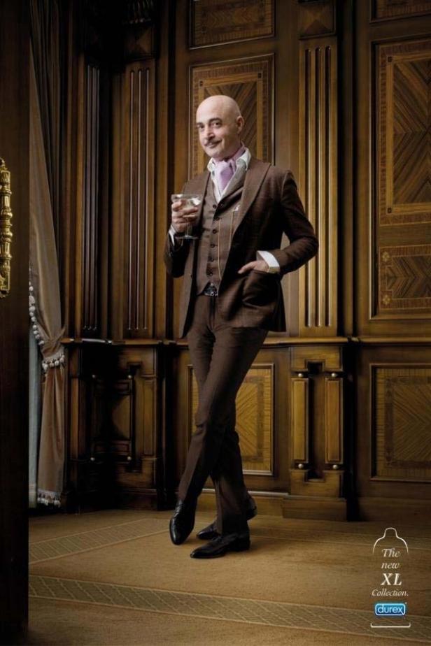Ein Mann im braunen Anzug posiert mit einem Drink in einem eleganten Raum.