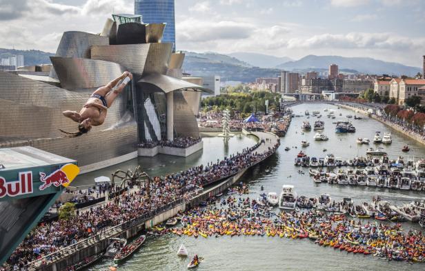 Ein Klippenspringer springt vor dem Guggenheim-Museum in Bilbao ins Wasser. Eine Menschenmenge und Boote beobachten das Spektakel.