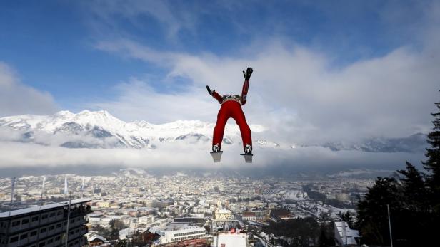 Ein Skispringer in rotem Anzug springt vor dem Panorama von Innsbruck und schneebedeckten Bergen.