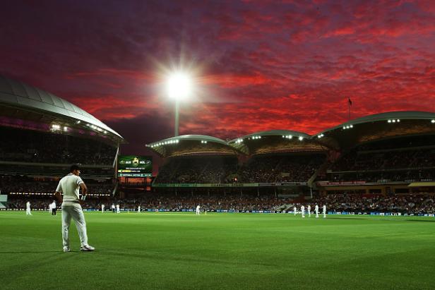 Ein Cricketspieler steht auf dem Feld vor einem Stadion im Abendrot.