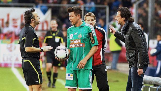 Ein Fußballspieler mit Ball diskutiert mit dem Schiedsrichter, während ein Trainer gestikuliert.