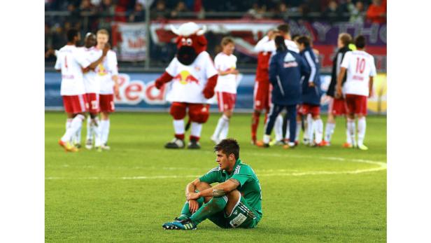 Ein enttäuschter Fußballspieler sitzt auf dem Rasen, im Hintergrund feiert das gegnerische Team.
