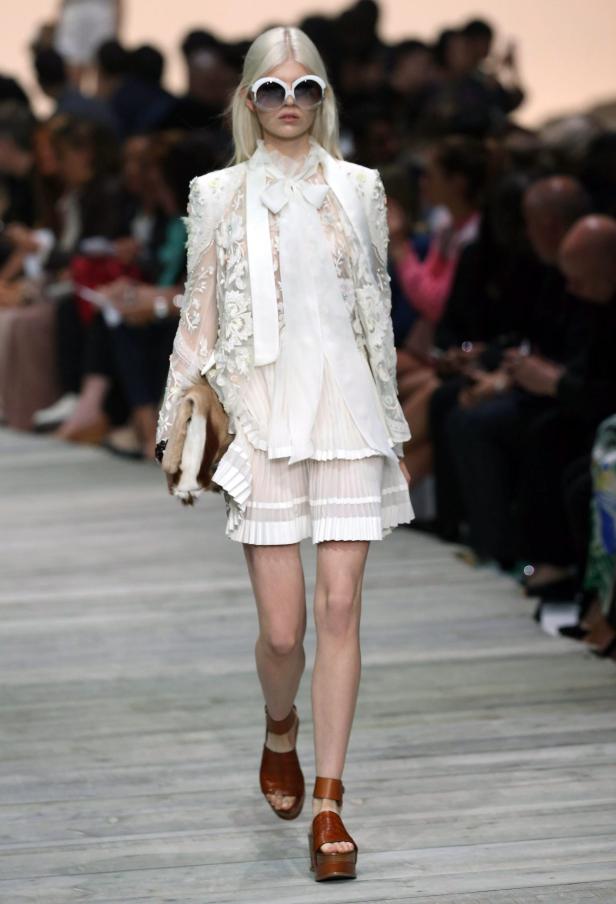 Ein Model in einem weißen Kleid mit Spitze und einer Fellhandtasche auf dem Laufsteg.